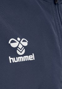 Marinblå jacka med en strukturerad tyg och en vit "hummel"-logotyp samt ett emblem ovanför som visar en bi-graphic.