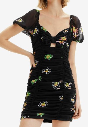 Robe mini noire froncée avec des manches bouffantes, un motif floral et une découpe nouée à l'avant sur la poitrine, portée par une personne avec la main sur la hanche.