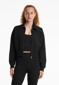 Mladá žena s kučeravými vlasmi, oblečená v čiernej skrátenej zipsovej bunde, čiernom crop top a čiernych legínach s vysokým pásom na bielom pozadí.