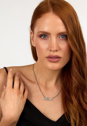 Collana d'argento con un design in corsivo "Zel", caratterizzata da accenti scintillanti, indossata da una persona con lunghi capelli rossi. Visibile una maglia scura.