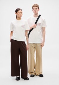 Valkoiset graafiset t-paidat, joissa logodetalji, yhdistetty leveälahkeisiin ruskeisiin ja beigen värisiin housuihin. Molemmilla mustat kengät ja ristiin kantaistavat laukut.