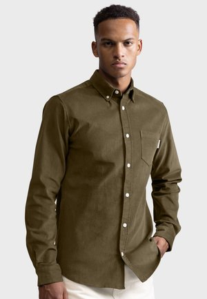 Chemise en velours côtelé vert olive avec un col boutonné, une poche poitrine unique et des boutons blancs ; présente une texture lisse et un ourlet droit.
