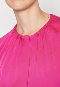 Fuchsia-Bluse mit Rundhalsausschnitt, die vertikale Falten und eine glatte, glänzende Textur aufweist. Keine sichtbaren Beschläge oder Muster.