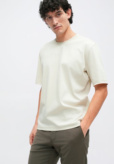 Világos bézs pamut póló rövid ujjal és kerek nyakkal, laza szabással és sima textúrával. Olívazöld nadrággal párosítva.