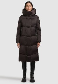 Musta eristetty toppatakki, jossa korkea kaulus, vetoketjulliset taskut ja tikattu kuvio. Pitkä pituus, sileä pinta ja istuvat hihat.