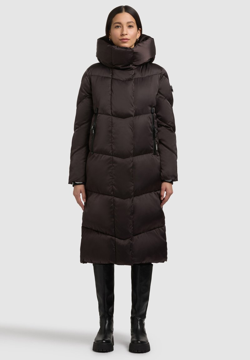 Musta eristetty toppatakki, jossa korkea kaulus, vetoketjulliset taskut ja tikattu kuvio. Pitkä pituus, sileä pinta ja istuvat hihat.