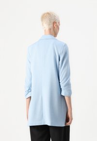 Ανοικτό μπλε μακρυμάνικο ενδύμα με υφή υφάσματος; διαθέτει γιακά, ρολαρισμένα μανίκια και ίσια γραμμή που φτάνει στο μέσο της μηρός.