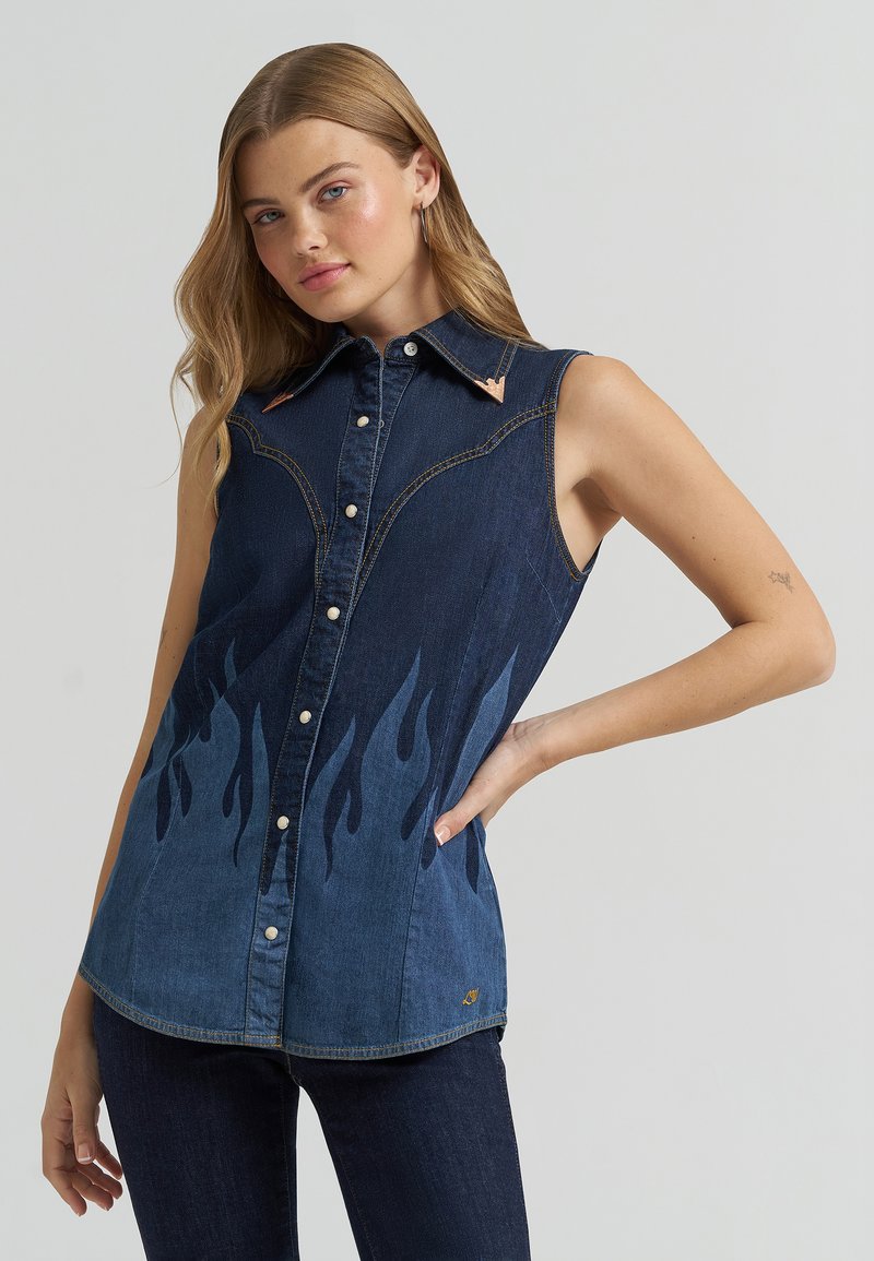 Γυναίκα με αμάνικο σκουρόχρωμο πουκάμισο από τζιν με σχέδιο φλόγας και μπρούτζινες άκρες γιακά, ποζάροντας με το ένα χέρι στη μέση μπροστά από απλό φόντο.