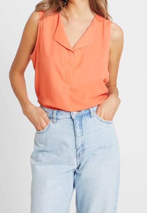Femme portant un chemisier orange sans manches avec col et un jean taille haute bleu clair, mains dans les poches, sur fond blanc.