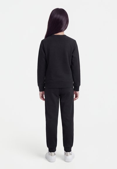 Bambino con capelli lunghi e scuri, indossa una felpa nera e pantaloni, in piedi di spalle, con scarpe da ginnastica bianche su sfondo semplice.