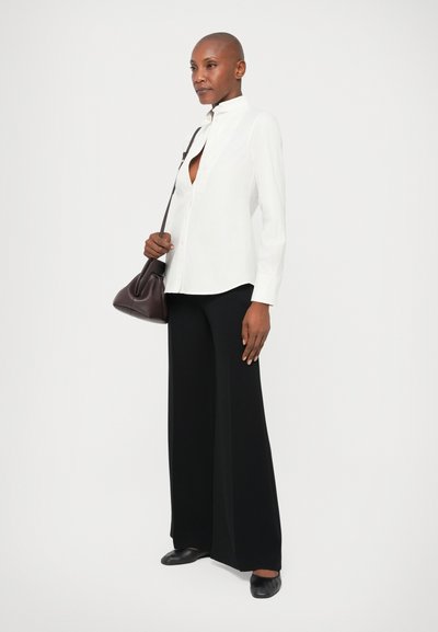 Biely košeľový tričko, voľné čierne nohavice a čierne slip-on topánky. Na neutrálnom pozadí. Tmavá kabelka vytvára kontrast.