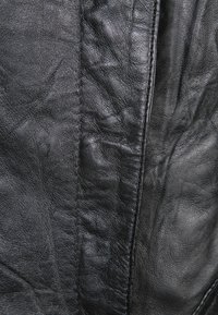 Schwarzes Ledergewebe mit einer strukturierten Oberfläche, die detaillierte Falten und Nähte zeigt. Das Material wirkt geschmeidig und gut abgenutzt.