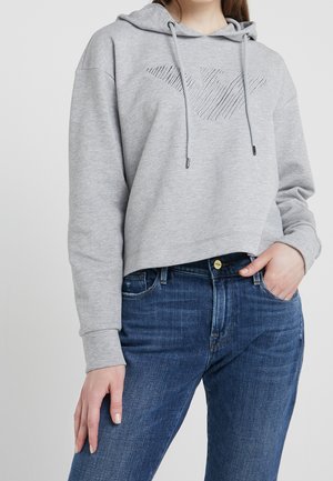 Osoba w szarej krótkiej bluzie z kapturem z naszytym geometrycznym logo i niebieskich dżinsach z wysokim stanem, z ręką w kieszeni.