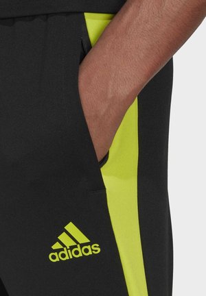 Main dans la poche d’un short Adidas noir avec une bande latérale jaune vif et le logo Adidas près de la poche.