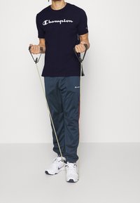 Tmavě modrá krátká tričko s bílým logem "Champion", šedé běžecké kalhoty s červenými bočními pruhy a bílé sportovní boty. V rukou drží odporové pásy.