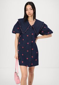 Τζιν φόρεμα με μεγάλο ρυ camo γιακά, κοντά φουσκωτά μανίκια και κεντημένη κόκκινη καρδιά. Το μοντέλο κρατάει μια ροζ τσάντα χειρός. Κουμπί μπροστινής κλεισίματος.