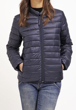 Kobieta ubrana w granatową pikowaną kurtkę zapinaną na zamek, z rękami w kieszeniach, w niebieskich dżinsach na białym tle.