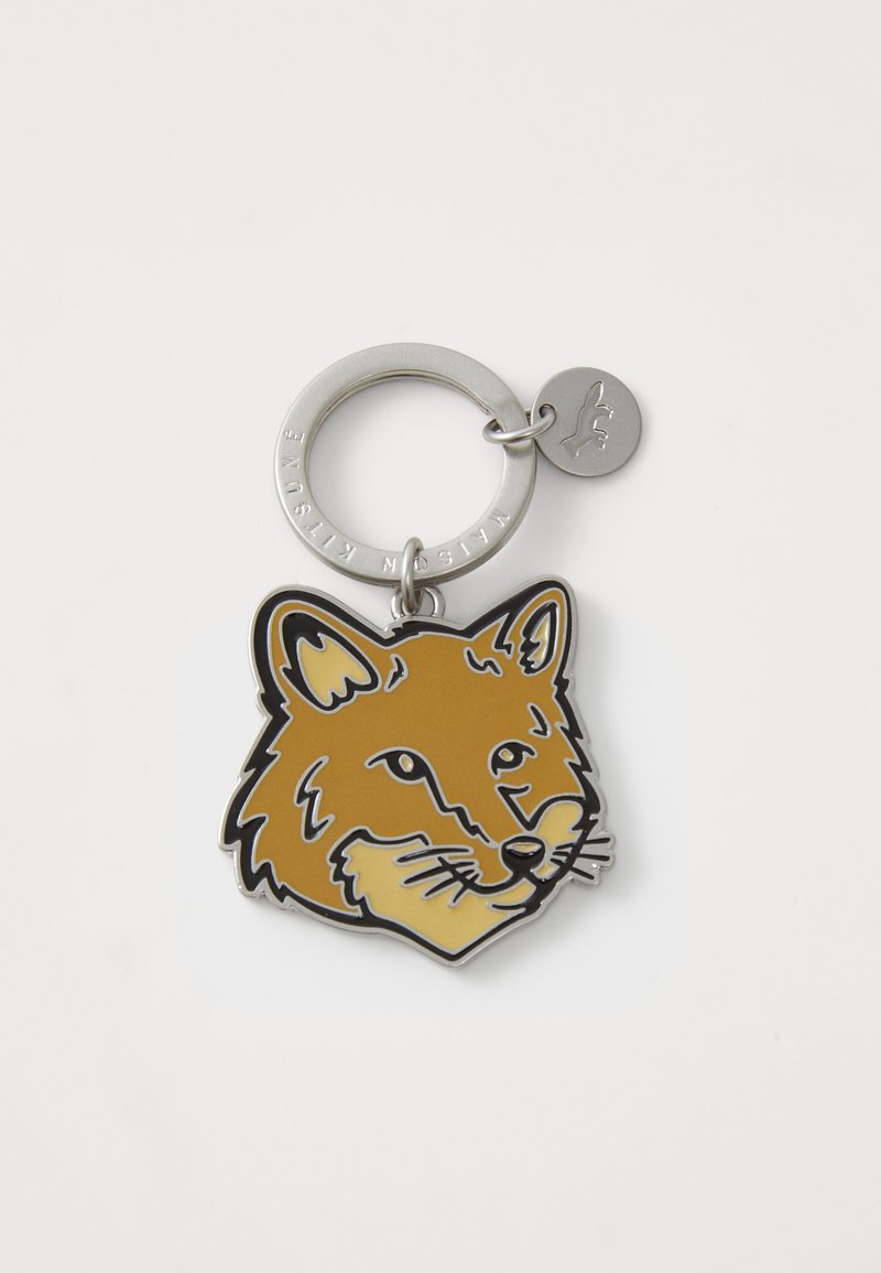 Porte-clés avec un visage de renard doré et marron, aux contours noirs, attaché à un anneau en métal argenté, avec une petite étiquette ronde.
