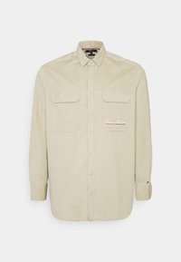 Beżowa koszula z długim rękawem z zapięciem na guziki, dwoma kieszeniami na piersi, haftowanym logo i podwiniętymi mankietami. Bawełniana tkanina, gładka w dotyku.