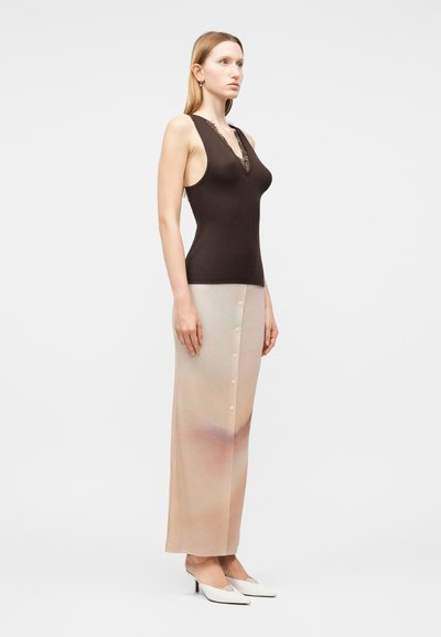 Γυναίκα σε προφίλ, φορώντας αμάνικη σκούρα καφέ μπλούζα με δαντέλα, μπεζ μακριά φούστα με κουμπιά και λευκές μυτερές γόβες.