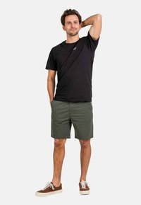 Camiseta de algodón negra de manga corta, combinada con pantalones cortos verde oliva. Zapatillas de ante marrón con suelas blancas completan el conjunto.