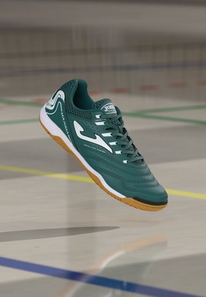 Scarpa da sport indoor turchese con logo bianco e suola in gomma, sospesa sopra il pavimento della palestra segnato con linee verdi, gialle, blu e rosse.