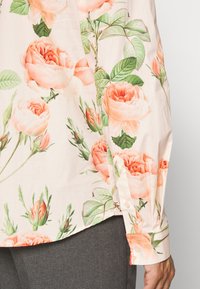 Blusa de manga larga con estampado floral en melocotón claro, con puños abotonados, combinada con pantalones gris oscuro, mostrando detalles de los lados y las mangas.