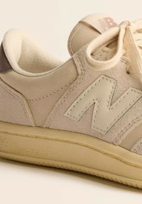 Beige suede and leather New Balance sneaker with stitched "N" logo, plush interior lining, laces tied in a bow, and textured rubber sole.
