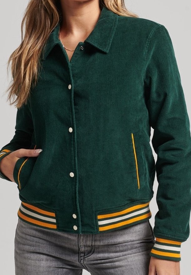 Femme portant une veste bomber en velours côtelé vert foncé avec des poignets et un ourlet côtelés rayés jaune et blanc, ainsi que des boutons-pression devant.