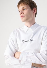 Balta ilga rankovė marškiniai su klasikinio kirpimo color, ant priekinės pusės yra juodas "Calvin Klein" užrašas. Lygus medvilnės audinys, sagčių rankogaliai.