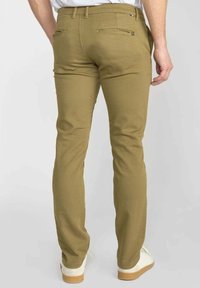 Man in slim-fit khaki broek met rits en knoopzakken aan de achterkant, gecombineerd met witte sneakers op een effen achtergrond.
