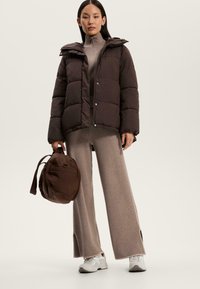 Giacca a vento marrone con cappuccio staccabile, maglia grigia a collo alto, pantaloni beige larghi con spacchi laterali e borsa marrone. Sneakers con suola testurizzata.