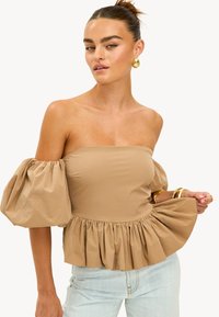 Haut beige à épaules dénudées avec des manches bouffantes, présentant un ourlet en peplum froncé. Tissu lisse avec une texture douce et des détails de conception minimaux.