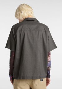 Chemise à manches courtes en tissu gris foncé avec un col, présentant une texture lisse et une coupe carrée. Étiquette visible sur l'ourlet inférieur.
