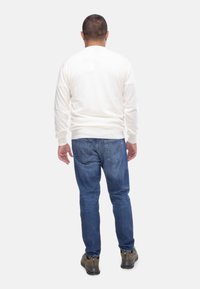 Maglione bianco a maglia con maniche lunghe, abbinato a jeans blu di denim. Modello indossa scarpe da trekking beige, visto da dietro.