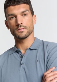 Lichtgrijze polo shirt met een gestructureerde stof, twee knopen bij de halslijn en een klein logo-detail op de linkerborst.