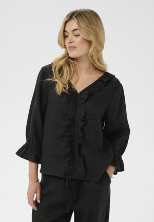 Mujer rubia con blusa negra con volantes en la parte delantera y las mangas, combinada con pantalones negros, de pie frente a un fondo liso.