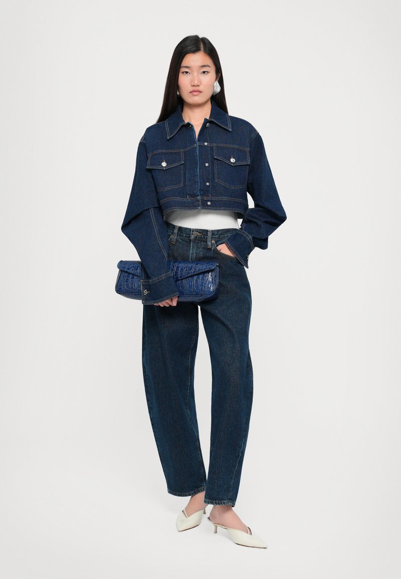 Cropped dark denim jacket with two front pockets, paired with loose dark jeans and a blue textured clutch. Model wears white heels.
