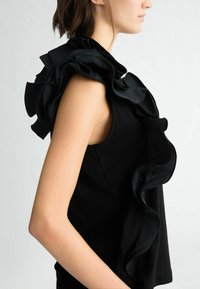 Top senza maniche nero con un colletto di raso arricciato. Il tessuto ha una texture liscia, mentre i volant aggiungono dimensione e movimento.