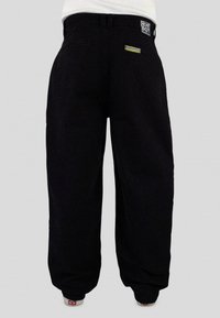 Black, loose-fitting pants with a textured fabric. Features a brand patch on the back and side pockets. Simple, straight-leg design.