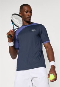 Camiseta de ténis de manga curta nas cores azul-marinho e roxo com logótipo, combinada com calções brancos. Modelo segura uma raquete de ténis e uma bola. Tecido texturizado.