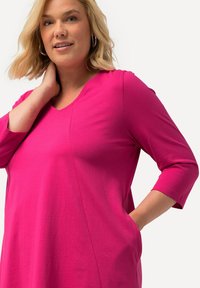 Blonde Frau mit Creolen, die ein leuchtend pinkes Oberteil mit 3/4-Ärmeln trägt, posiert mit einer Hand am Hals und der anderen in der Tasche.