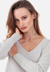 Hellgrauer Pullover mit langen Ärmeln und V-Ausschnitt, mit weicher Textur und eng anliegenden Ärmeln, getragen mit einer Hand am Hals.