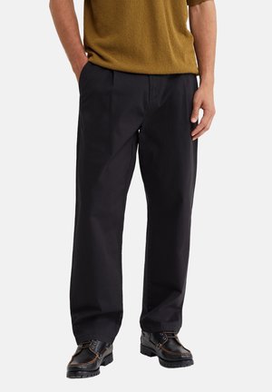 Homme portant un pantalon noir droit, des chaussures noires à lacets et un pull marron à manches courtes, les mains dans les poches.