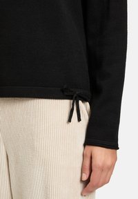 Schwarzer Pullover mit langen Ärmeln, ausgestattet mit einem strukturierten Stoff und einem verstellbaren Kordelzug am Saum. Kombiniert mit hellbeigen Cordhosen.