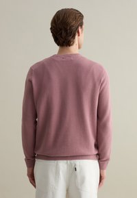 Pink knitted sweater with a textured pattern, crew neckline, and ribbed cuffs. Worn with white pants, showing a relaxed fit and casual design.