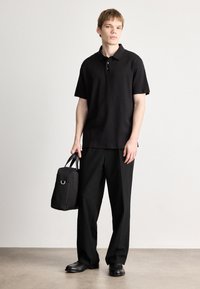 Camisa polo preta de manga curta, combinada com calças largas pretas. O modelo segura uma bolsa preta com alça superior. Tecido liso, design minimalista.