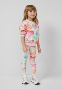 Junges Mädchen in buntem Sweatshirt mit Herzmuster und Leggings, trägt weiße Sneakers vor einfachem grauem Hintergrund.