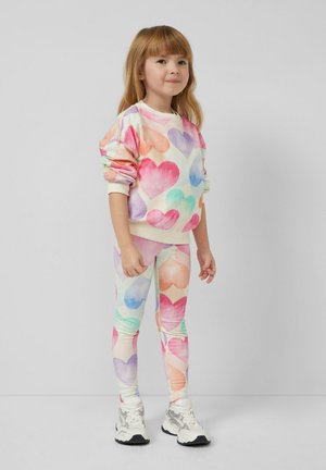 Jong meisje in een kleurrijke trui met hartjespatroon en legging, met witte sneakers, staand tegen een effen grijze achtergrond.