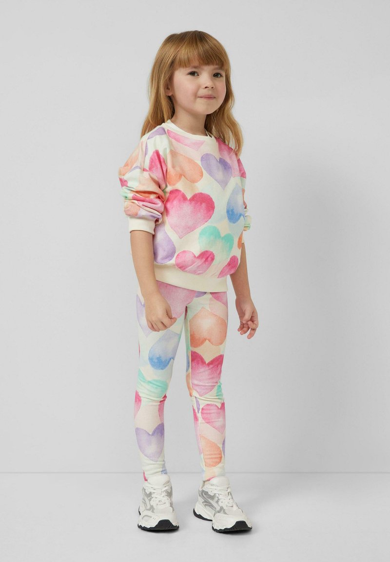 Junges Mädchen in buntem Sweatshirt mit Herzmuster und Leggings, trägt weiße Sneakers vor einfachem grauem Hintergrund.